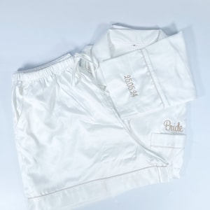 May include: White satin pajama set with shorts and a button-down shirt. The shirt cuff is embroidered with the date "25.05.24" in gold thread, and the word "Bride" is embroidered on the pocket. The set includes a drawstring waist.