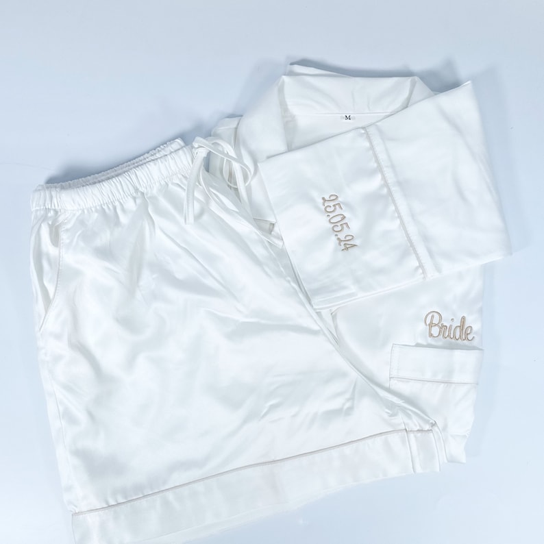 May include: White satin pajama set with shorts and a button-down shirt. The shirt cuff is embroidered with the date "25.05.24" in gold thread, and the word "Bride" is embroidered on the pocket. The set includes a drawstring waist.