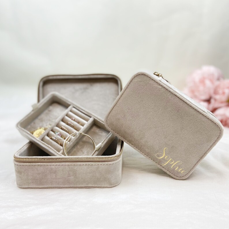 May include: A gray velvet jewelry box with a zipper closure. The box is open and has a smaller compartment inside for rings and earrings. The box is personalized with the name "Sophia" in gold lettering.