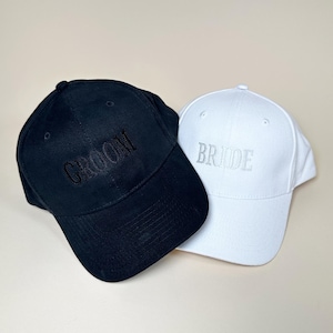 May include: Two baseball caps, one black with the word "Groom" embroidered in black, and one white with the word "Bride" embroidered in silver.