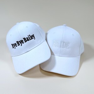 May include: Two white baseball caps. One cap says "Bye Bye Bailey" in black lettering. The other cap says "Bride" in white lettering.