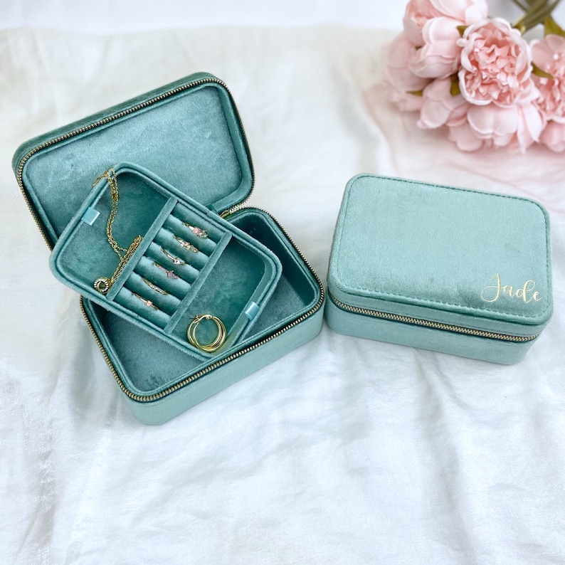 May include: Two teal velvet jewelry boxes with gold zippers. The larger box is open and has a compartment for necklaces and earrings. The smaller box is closed and has the name "Jade" written on it in gold lettering.