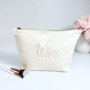 May include: A white canvas makeup bag with a zipper closure. The bag has the words "Wifey Est. 2023" embroidered on the front in a gold thread.