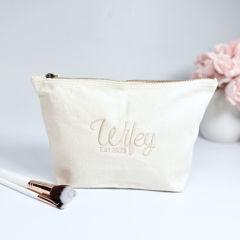 May include: A white canvas makeup bag with a zipper closure. The bag has the words "Wifey Est. 2023" embroidered on the front in a gold thread.