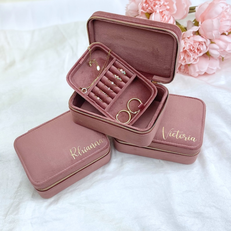 May include: Three pink velvet jewelry boxes with gold zippers. The boxes are stacked on top of each other. The top box is open and shows a compartment for rings and earrings. The middle box has the name "Rhianna" written on it in gold lettering. The bottom box has the name "Victoria" written on it in gold lettering.
