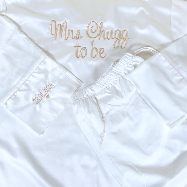 May include: White satin pajama set with gold embroidery. The top reads "Mrs Chugg to be" and the sleeve cuff has "23.01.2024" embroidered. The set includes a long-sleeved top and matching pants.