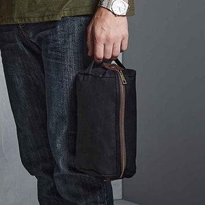 May include: A black canvas wash bag with a brown leather handle and zip. The bag is approximately 8 inches tall and 5 inches wide.