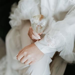 May include: A white robe with ruffled sleeves, holding a champagne glass. The sleeve features embroidered text and a heart. The robe is made of a soft, flowing fabric, perfect for bridal wear or a special occasion.