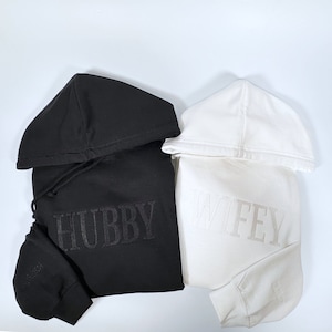 May include: Two hooded sweatshirts, one black and one white. The black sweatshirt has "HUBBY" embroidered on the front, and the white sweatshirt has "WIFEY" embroidered on the front. Both have drawstrings and a small text on the sleeve.