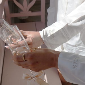 May include: Two champagne flutes being filled with a sparkling beverage. The flutes have a decorative bow and a ring with the word "DIOR". The sleeves of a white shirt have the numbers "07.04.16" and "07.04.26" embroidered on them.