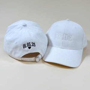 May include: Two white baseball caps, one with the word "BRIDE" embroidered on the front and the other with the date "01.05.25" and a heart embroidered on the back.