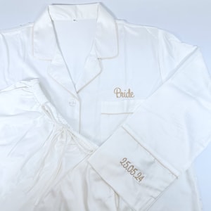 May include: Ivory satin pajama set with light tan trim. The top has "Bride" embroidered on the pocket, and the cuff of the sleeve has "25.05.24" embroidered. The pajama top has a button-down front and a notched collar. The pants have a drawstring waist.