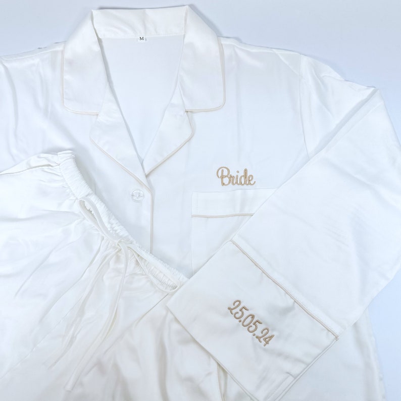 May include: Ivory satin pajama set with light tan trim. The top has "Bride" embroidered on the pocket, and the cuff of the sleeve has "25.05.24" embroidered. The pajama top has a button-down front and a notched collar. The pants have a drawstring waist.