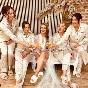 May include: A group of five women in matching beige satin pyjamas with black piping, raising champagne glasses. The bride is in a white lace robe and veil. The scene is a bridal party, with a slatted wooden wall and floral display behind them.
