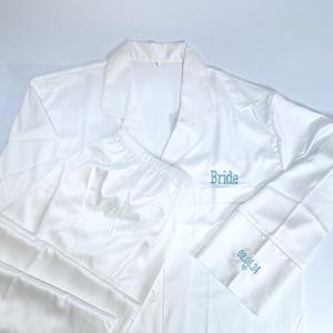 May include: White satin pajama set with the word "Bride" embroidered in teal on the chest pocket. The cuff of the long-sleeved shirt has the date "08.06.24" embroidered in teal. The set includes a button-down shirt and matching pants.