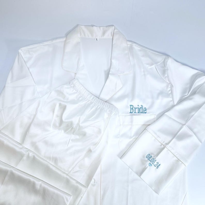 May include: White satin pajama set with the word "Bride" embroidered in teal on the chest pocket. The cuff of the long-sleeved shirt has the date "08.06.24" embroidered in teal. The set includes a button-down shirt and matching pants.