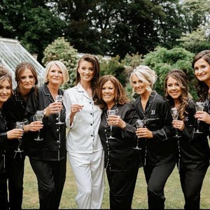 May include: A group of eight individuals wearing matching button-down pyjama sets with long sleeves and collars. Seven individuals wear black pyjama sets with white piping, while one individual in the centre wears a white pyjama set with black piping.