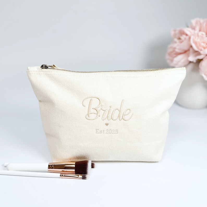 May include: A white canvas makeup bag with a gold zipper. The bag has the word "Bride" embroidered on it in a cursive font, along with a small heart and the text "Est 2025".
