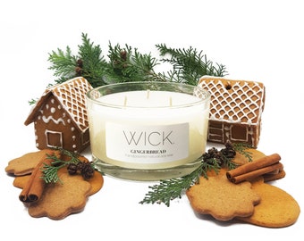 May include: A large, clear glass candle with the text "WICK GINGERBREAD" sits among gingerbread cookies, cinnamon sticks, and miniature gingerbread houses. Greenery adds a festive touch to the holiday-themed arrangement.