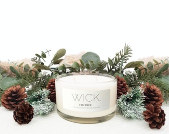 May include: A large, clear glass candle with four wicks, labeled "WICK. FIR TREE." The candle is surrounded by pinecones, greenery, and decorative foliage. The candle is a creamy white color and is hand-poured with natural soy wax.