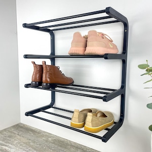 May include: A black metal shoe rack with three tiers, holding various shoes. The top shelf displays pink sneakers, the middle shelf has brown leather boots, and the bottom shelf holds tan slip-on shoes. The rack is mounted on a white wall.