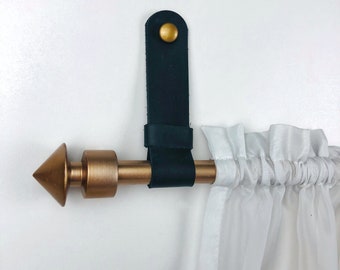 Könnte beinhalten: Ein schwarzer Leder-Vorhang-Halter mit einem goldenen Knopf, befestigt an einer goldenen Metall-Vorhangstange mit einem spitzen Abschluss.