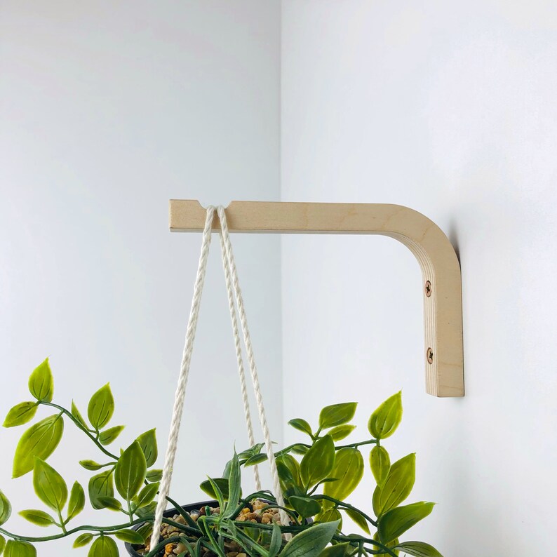 Puede incluir: Un soporte de pared para plantas hecho de madera clara con un dise&ntilde;o curvo, fijado con dos tornillos visibles. Una cuerda blanca trenzada cuelga del extremo con muesca del soporte, sosteniendo una planta verde en una maceta.