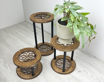 Puede incluir: Tres soportes de plantas de madera con acabado marrón oscuro y patas de metal negro. Cada soporte presenta un diseño floral decorativo en la parte superior e inferior. Un soporte sostiene una planta en maceta con follaje verde.