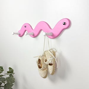 Könnte beinhalten: Eine rosafarbene Wandgarderobe in Wurmform mit gewelltem Körper und drei weißen Haken. Die Garderobe hat zwei kleine, runde, silberfarbene Augen. Ein Paar beigefarbene Schlupfschuhe mit Spitzenstruktur und weißen Bändern hängt an einem Haken.