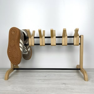 May include: A wooden shoe rack with black metal bars and six shoe hooks. The rack is made of light wood and has a simple, modern design. A pair of brown and white sneakers are hanging on the rack.