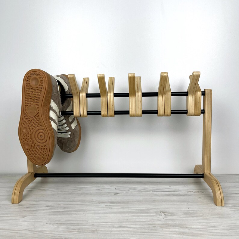 May include: A wooden shoe rack with black metal bars and six shoe hooks. The rack is made of light wood and has a simple, modern design. A pair of brown and white sneakers are hanging on the rack.