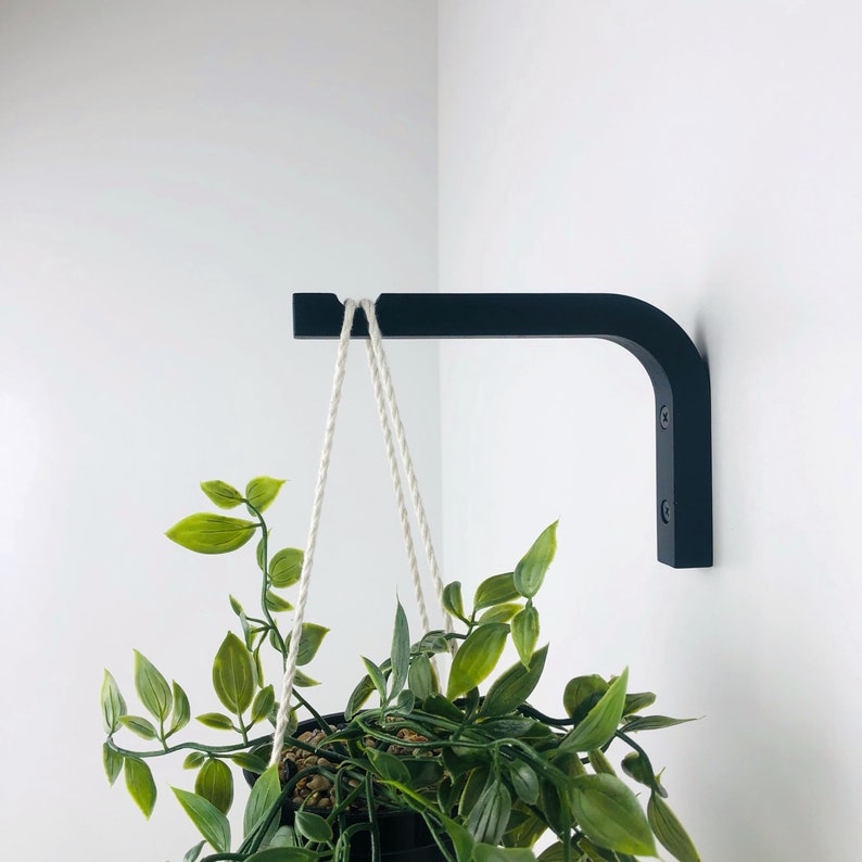 Puede incluir: Un soporte de pared en forma de L de color negro mate montado en una pared blanca, con un extremo con muesca para asegurar un colgador de cuerda blanca. Una planta de hojas verdes en una maceta negra cuelga del soporte con la cuerda.