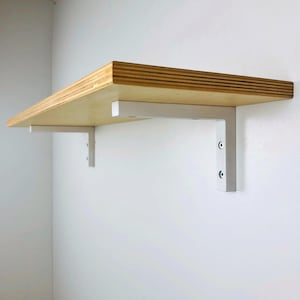 May include: A wooden shelf with white metal brackets. The shelf is made of plywood and has a natural wood finish. The brackets are attached to the underside of the shelf and are secured to the wall.
