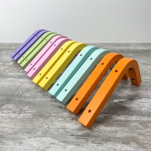 Pode incluir: Uma coleção de suportes de madeira curvos e coloridos em tons pastel. Cada suporte tem uma forma de ângulo reto com orifícios pré-perfurados para montagem. As cores incluem lavanda, verde, rosa, amarelo, azul claro e laranja.