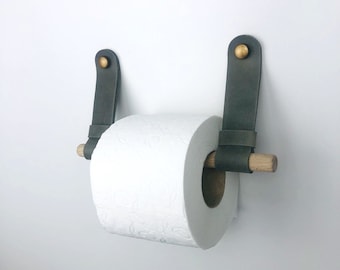 May include: A toilet paper holder featuring two dark gray leather straps secured to the wall with brass-colored metal studs. A light-colored wooden dowel passes through the loops of the leather straps to support a roll of white toilet paper.