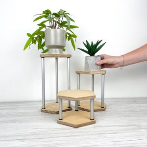 May include: Three tiered display stands featuring hexagonal light wood platforms supported by silver metal cylindrical legs. The stands vary in height, with one tall, one medium, and one short unit, designed to hold potted plants or decorative items.