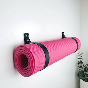 Pu&ograve; includere: Un tappetino da yoga rosa &egrave; arrotolato e fissato con un cinturino in pelle nera. Il tappetino &egrave; appeso a una cinghia montata a parete.