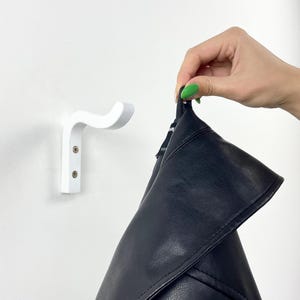 Pode incluir: Um gancho de parede branco com um design curvo. O gancho é feito de madeira e é fixado à parede com dois parafusos. Uma jaqueta preta está sendo segurada pelo gancho.