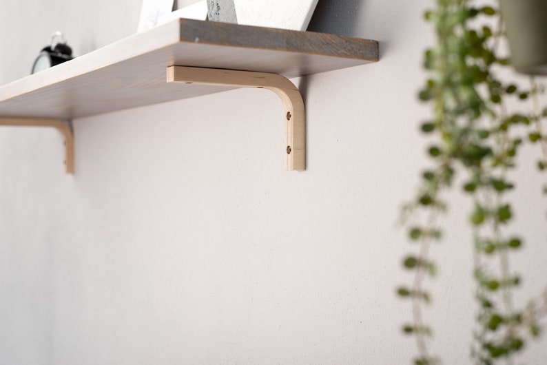 May include: A wooden shelf bracket with a curved edge, mounted to a white wall. The bracket is made of light wood and has a natural finish.