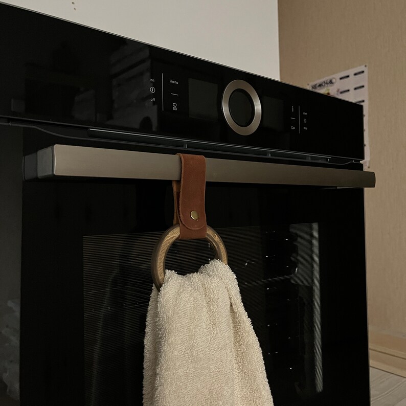 May include: A brown leather towel ring with a wooden ring hangs on a black oven door handle. A white towel is hanging on the ring.