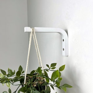 Puede incluir: Un soporte de pared blanco en forma de L montado en la pared, con una muesca en el extremo para colgar objetos. Una planta en maceta con hojas verdes cuelga del soporte mediante una cuerda trenzada de color claro, justo debajo del gancho.