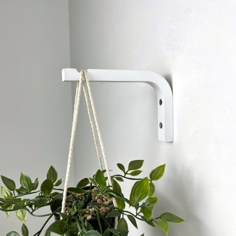 Puede incluir: Un soporte de pared blanco en forma de L montado en la pared, con una muesca en el extremo para colgar objetos. Una planta en maceta con hojas verdes cuelga del soporte mediante una cuerda trenzada de color claro, justo debajo del gancho.