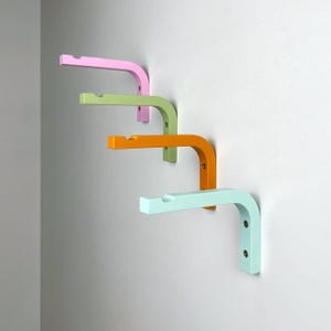 May include: Four wall-mounted shelf brackets in different pastel colours: light pink, light green, orange, and light blue. The brackets are made of wood and have a simple, modern design.
