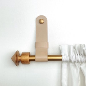 May include: A beige leather curtain tieback with a gold-colored rod and finial. The tieback is attached to the wall with a gold-colored button. White curtains are visible on the right side of the image.