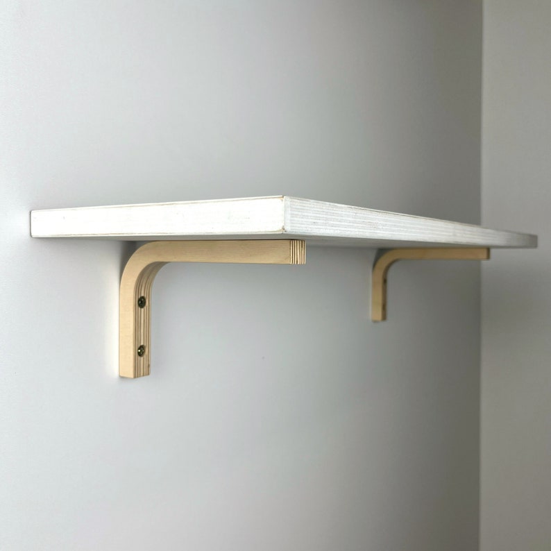 May include: White wooden shelf with two light brown wooden brackets. The shelf is mounted to a white wall.