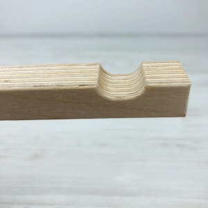Puede incluir: Una tira de madera clara hecha de contrachapado laminado, con una muesca semicircular lisa cortada en el borde superior. La veta de la madera es visible a lo largo de las capas horizontales, mostrando los tonos naturales beige y arena.