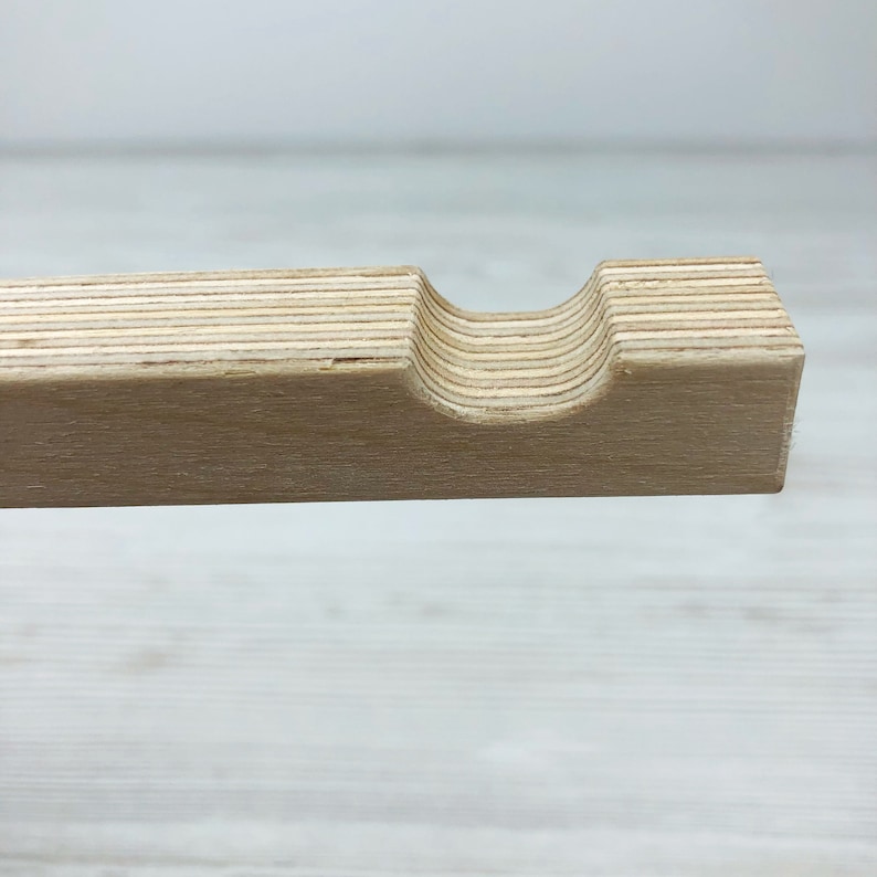 Puede incluir: Una tira de madera clara hecha de contrachapado laminado, con una muesca semicircular lisa cortada en el borde superior. La veta de la madera es visible a lo largo de las capas horizontales, mostrando los tonos naturales beige y arena.