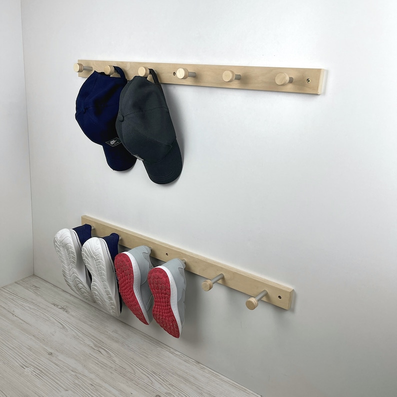 May include: A wooden wall-mounted rack with hooks for hanging hats and shoes. Two baseball caps hang on the top rack, one blue and one black. Three pairs of shoes hang on the bottom rack, one pair white, one pair red, and one pair grey.