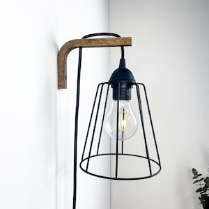 May include: A contemporary wall lamp featuring a black metal cage shade and a clear lightbulb. The lamp is mounted on a wooden arm and has a black cord. The design is minimalist and industrial, suitable for various interiors.