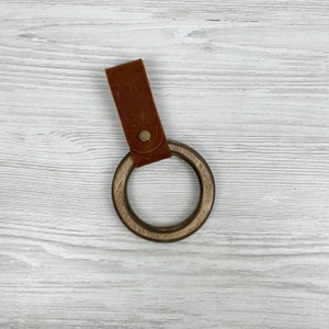 May include: A wooden ring with a brown leather strap. The ring is made of dark wood and has a smooth finish. The strap is attached to the ring with a metal rivet.